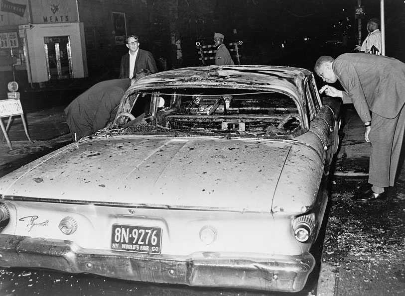 Detective examines burned out police car following rioting. Bedford-Stuyvesant, Brooklyn, 1964. (Stanley Wolfson / Library of Congress)